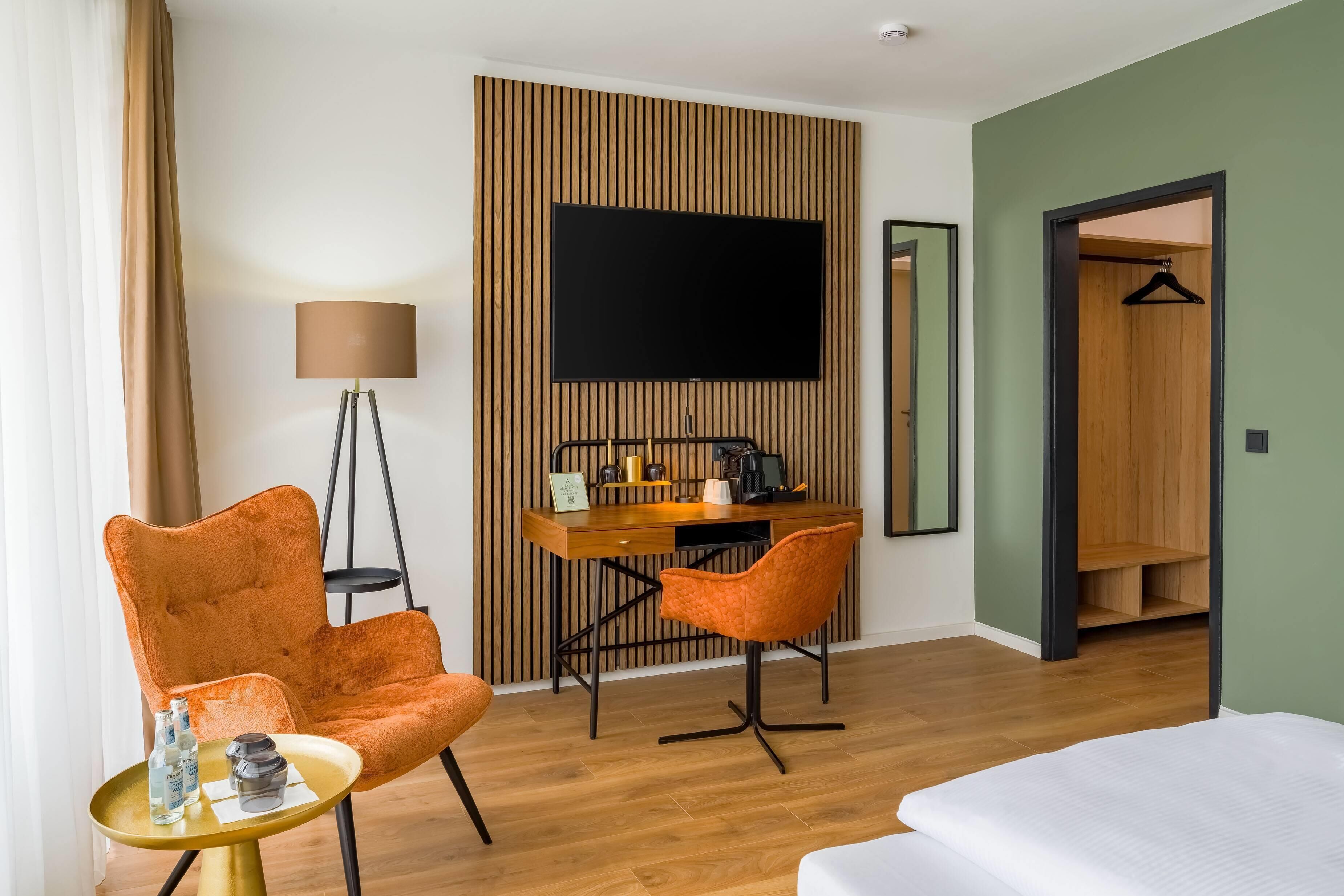 A hotel room with a wooden desk, two orange chairs, a bed, a TV, and a wardrobe.