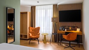 A hotel room with a bed, two orange chairs, a desk with a computer, a TV, and a wardrobe. - Aspire Palais Recklinghausen,Trademark Collection by Wyndham (Recklinghausen)