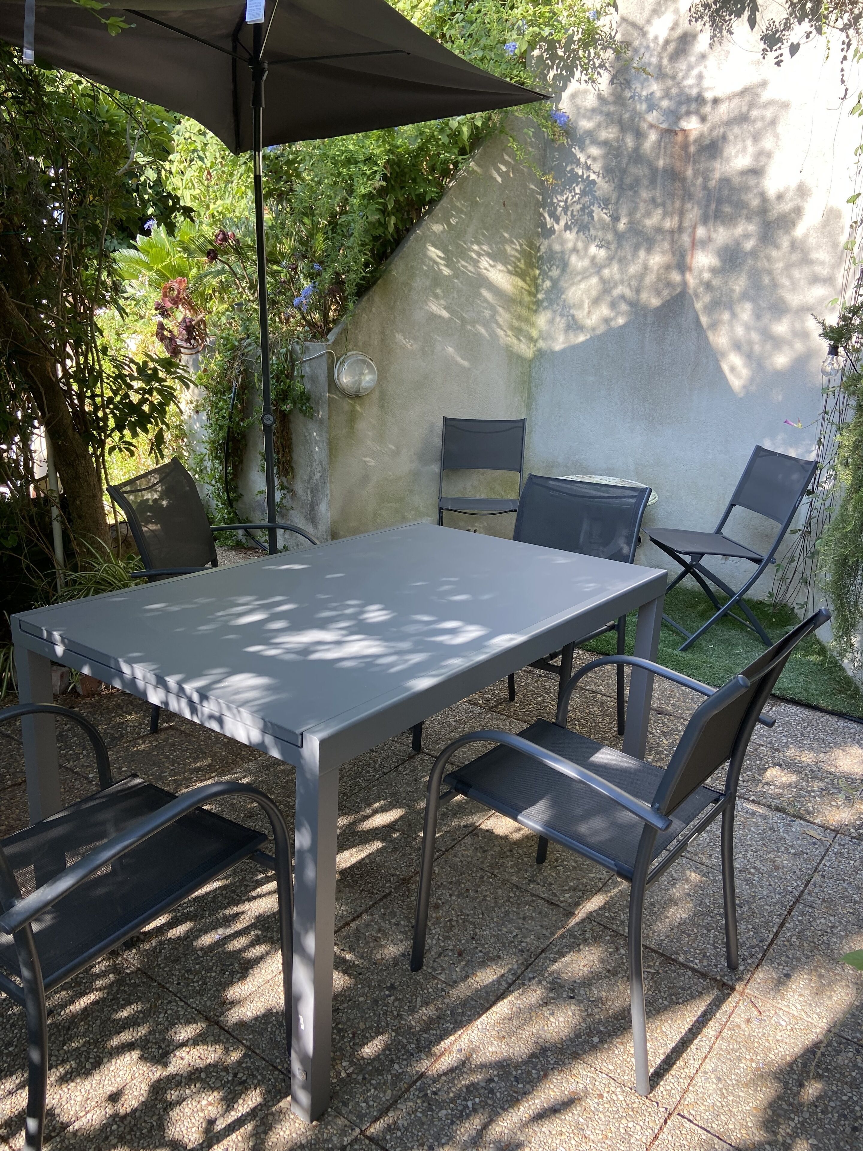 An outdoor patio with a table and four chairs, surrounded by greenery and a stone wall.
