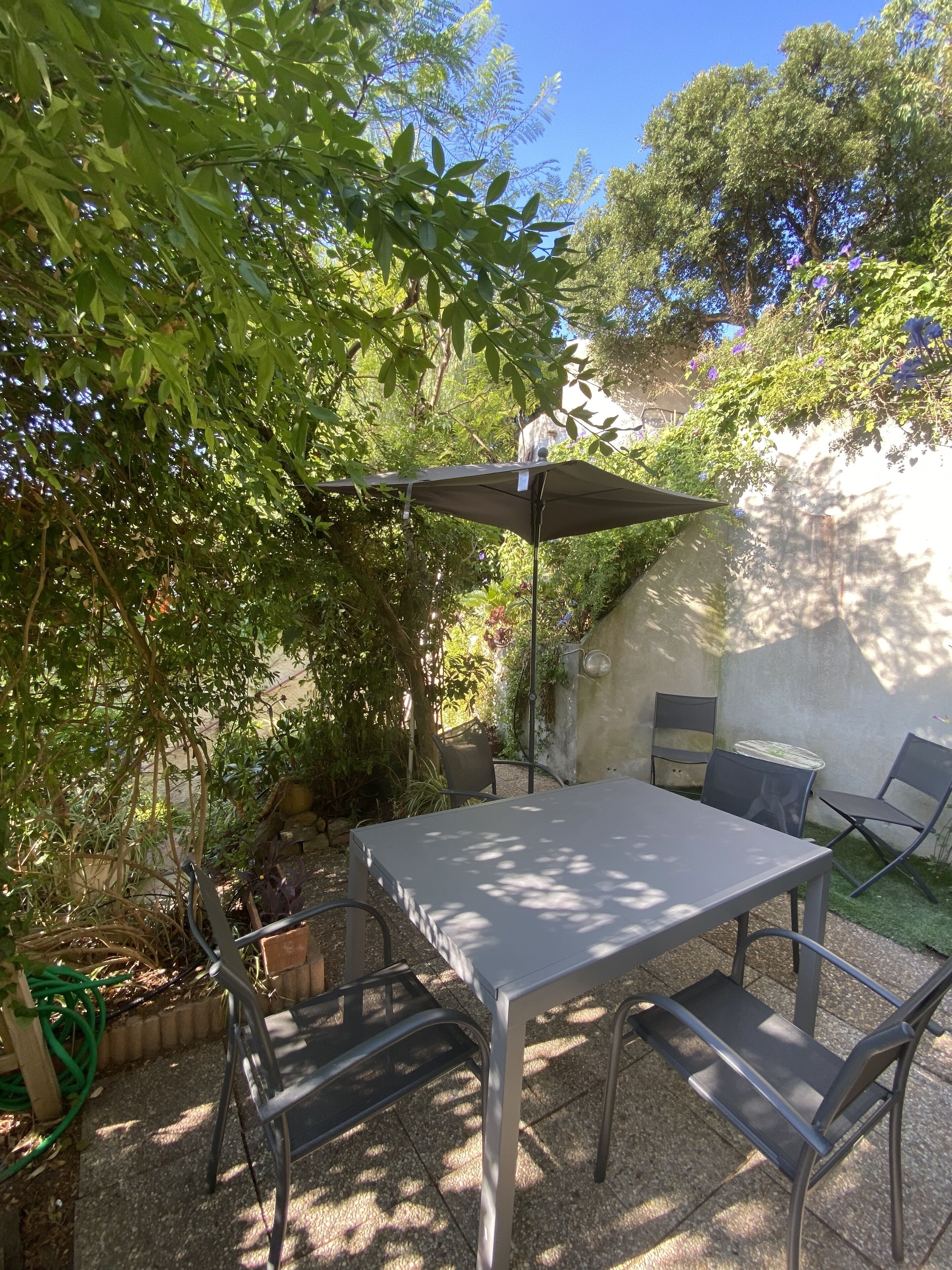 Un patio al aire libre con una mesa y sillas, rodeado de follaje y una pared.