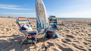 Escena de platja amb dues cadires de bany estriades, una taula de surf i una bossa a la sorra.