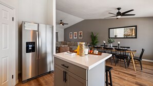 A modern kitchen with a stainless steel refrigerator, a white countertop, and a dining area with a wooden table and chairs. - Montego by AvantStay | Private Yard w/ Pool & Hot Tub (Indio)