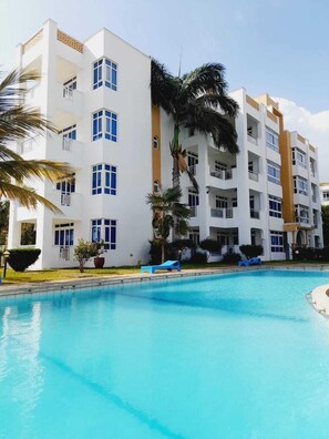 A multi-story hotel with a swimming pool, palm trees, and a clear sky. - Lux Suites Dofils Almasi Apartments (Mombasa)