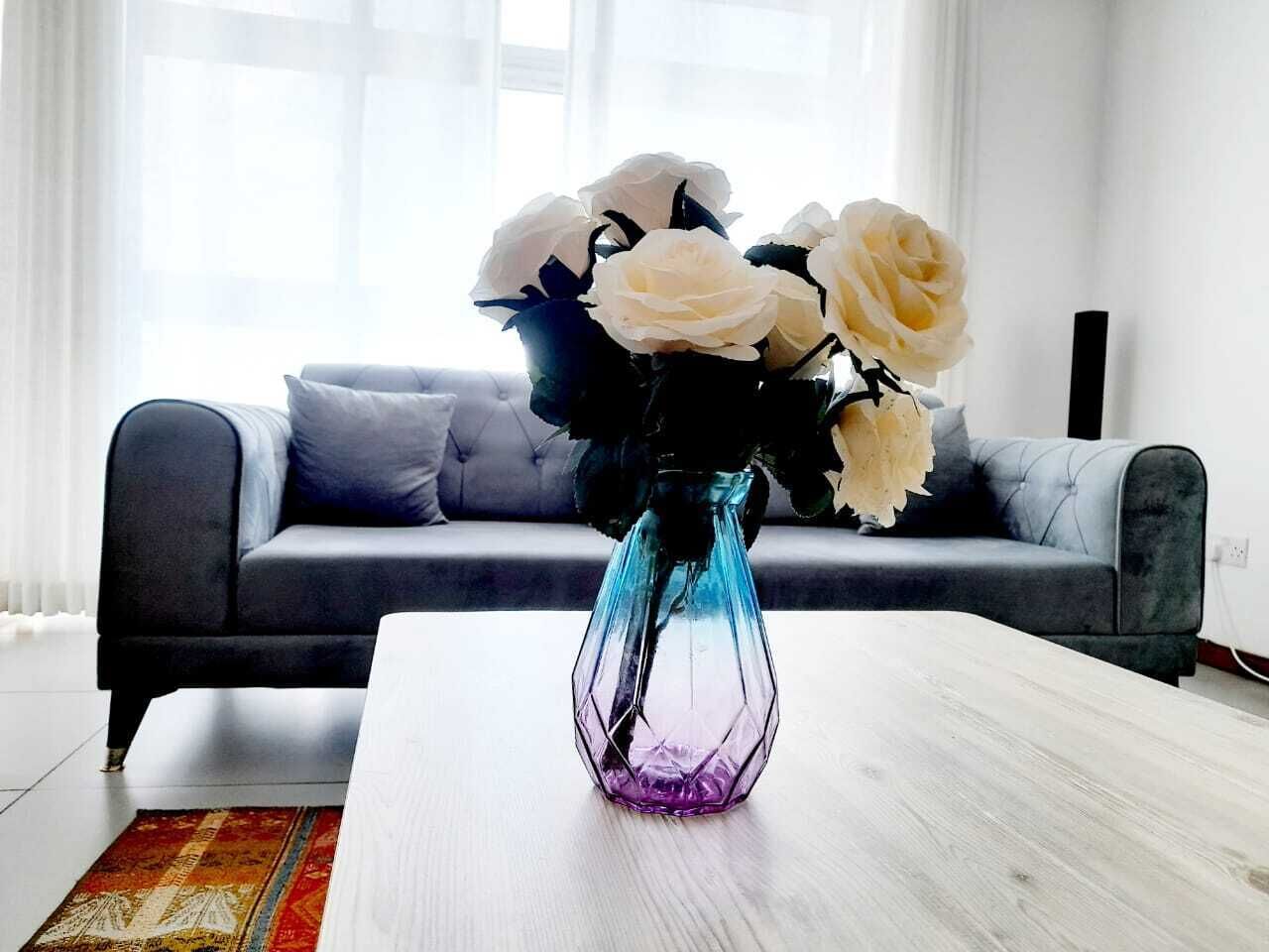 Uma sala de estar com um sofá cinzento, uma mesa de centro branca e um vaso com flores brancas.