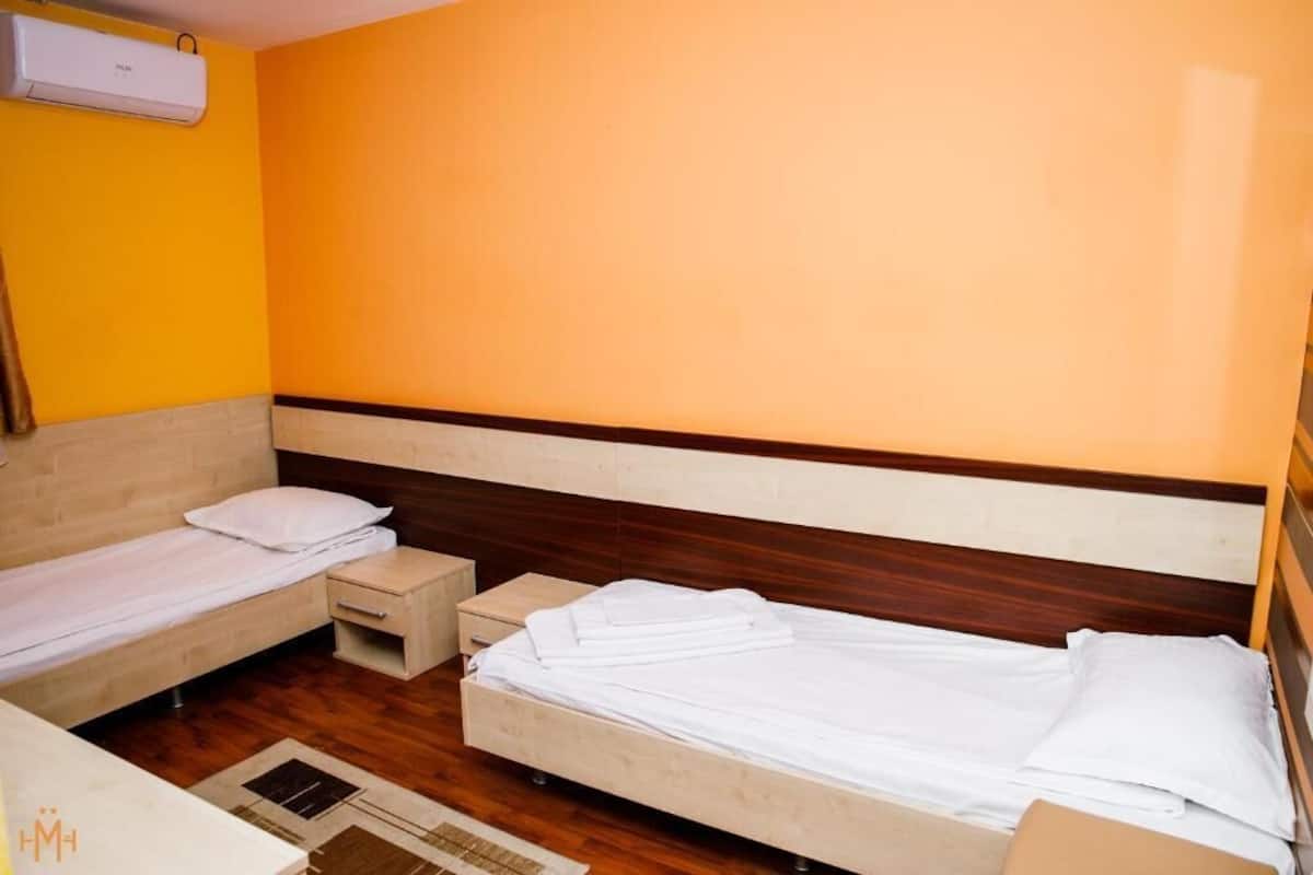 Hotellihuone, jossa on kaksi sänkyä, puulattia, pieni yöpöytä ja seinälle asennettu ilmastointilaite.