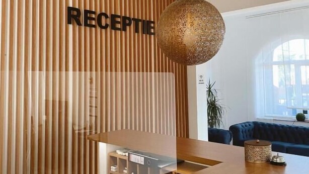 Un área de recepción con un muro de listones de madera, un mostrador de recepción, una lámpara colgante esférica y un sofá tapizado azul al fondo.
