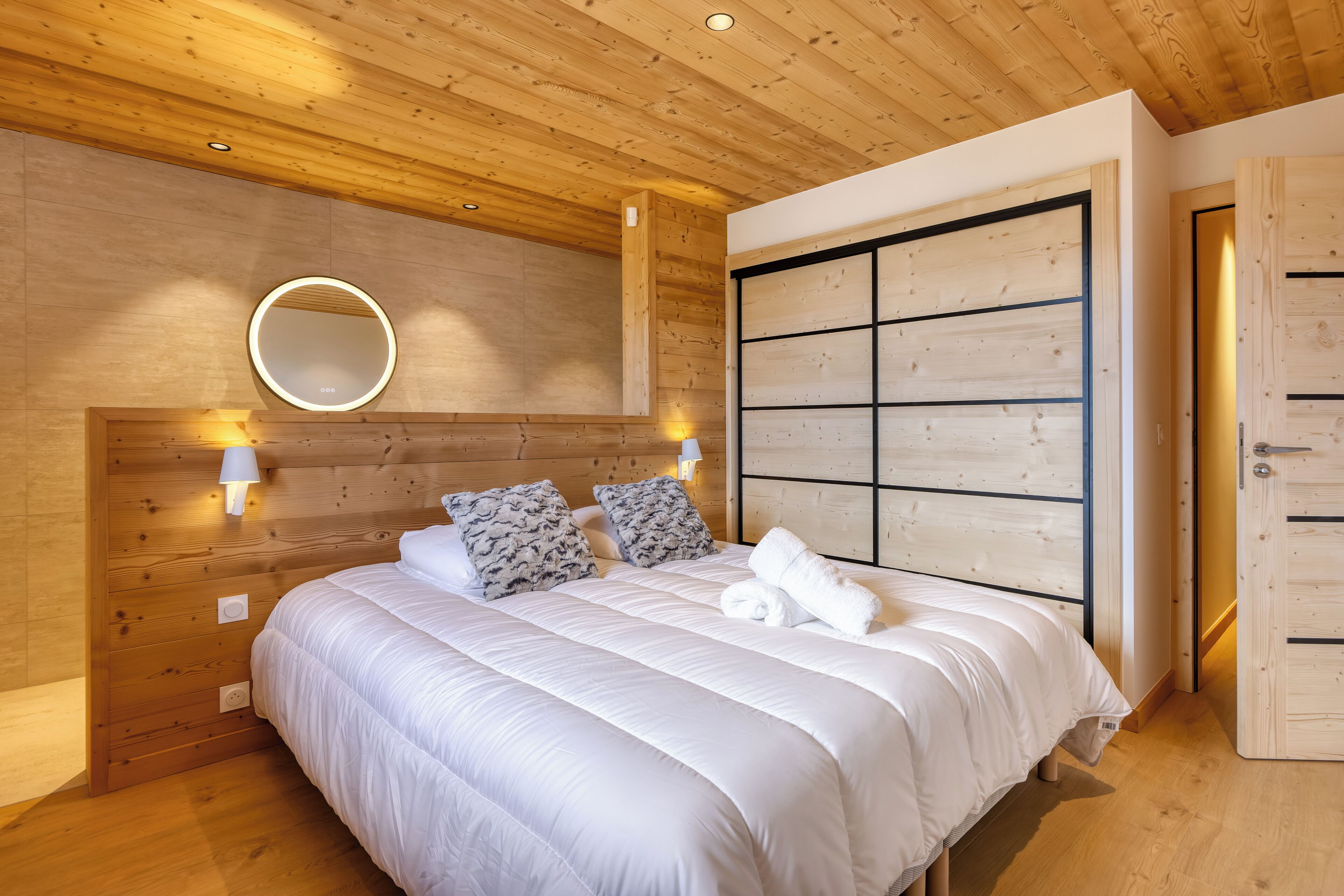 Een slaapkamer met een houten plafond, een groot bed met wit beddengoed, twee wandlampen en een houten garderobe.
