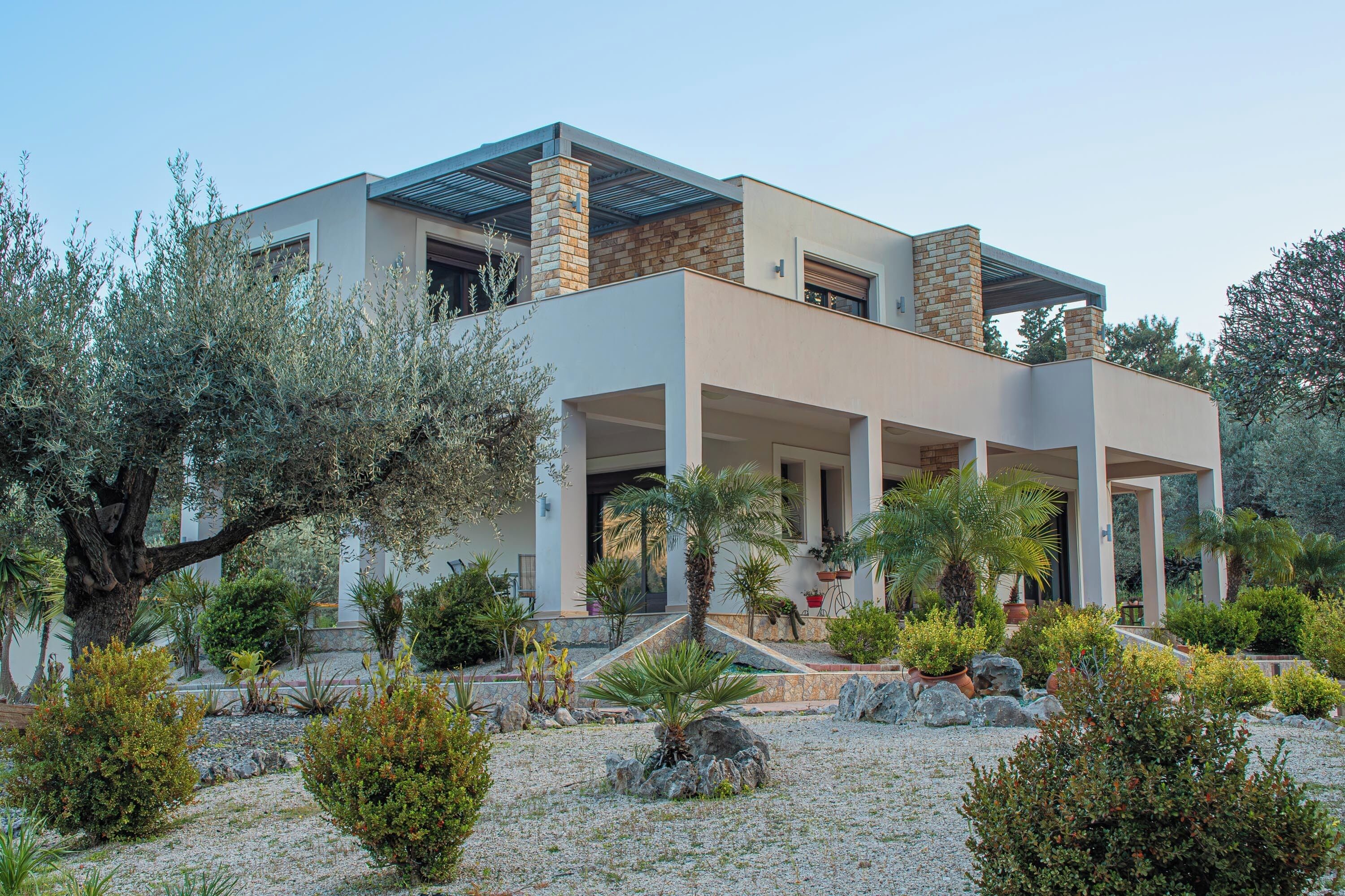 A modern two-story house with a covered patio, surrounded by a landscaped garden with various plants and a large tree.