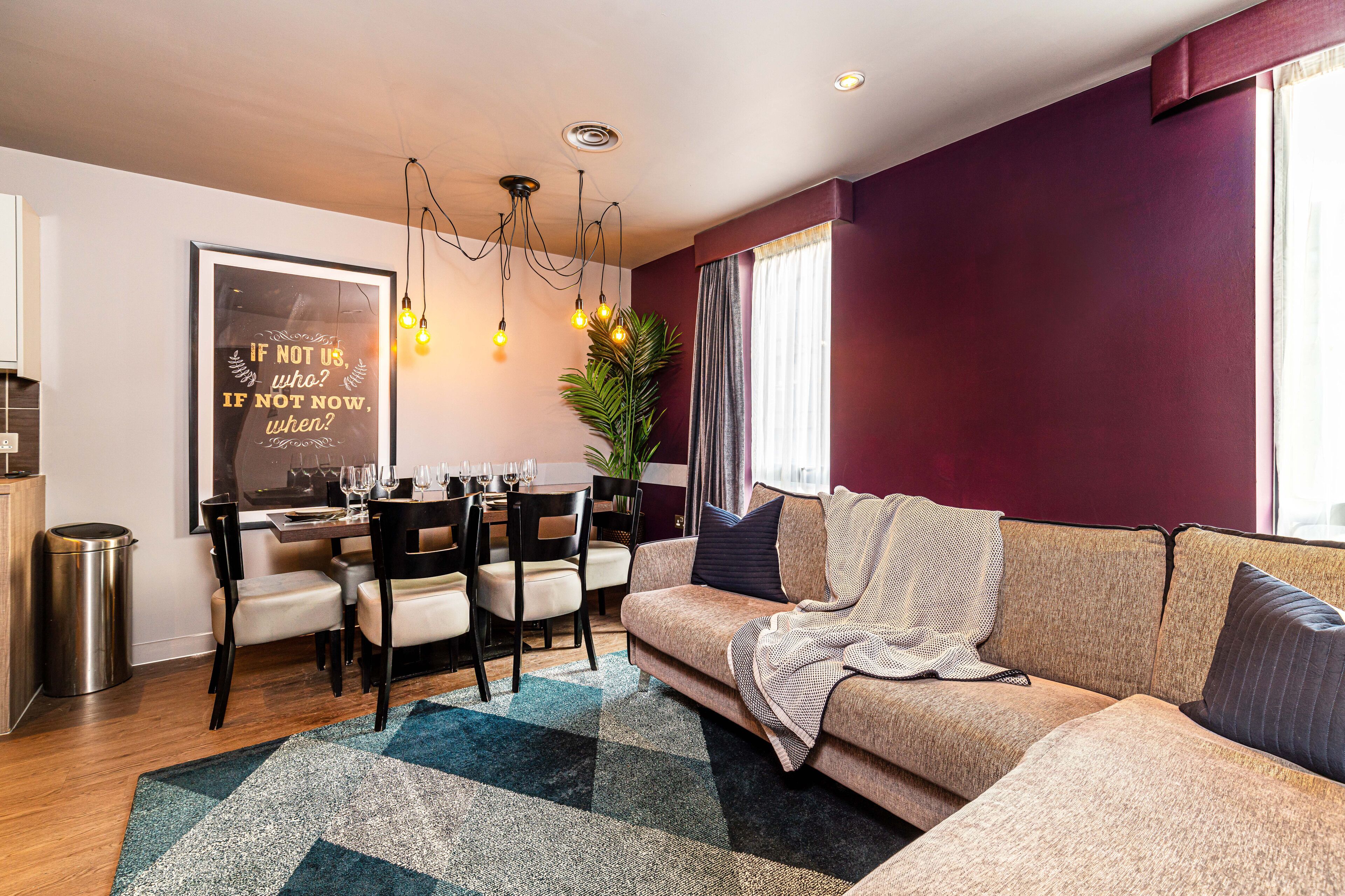 A modern living room with a sofa, dining table, and chairs. There is a framed quote on the wall and a plant in the corner.