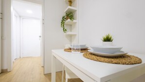 Una mesa de comedor blanca con un mantel tejido, un tazón y un vaso. Una planta en maceta está sobre una estantería blanca.