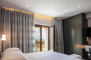 A modern hotel room with a large bed, a desk, and a view of the sea through a sliding door.