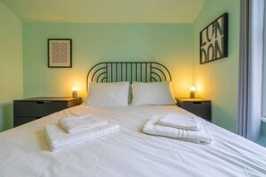 A neatly made bed with white linens and towels, two bedside tables with lamps, a framed picture on the wall, and a sign that reads "LONDON".