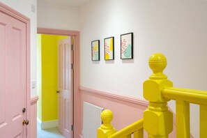 A hallway with a pink door, a yellow railing, and three framed artworks on the wall.