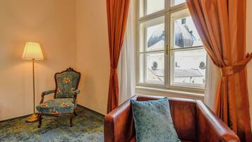 Ein Zimmer mit einem blauen, floralen Ohrensessel, einem braunen Ledersofa mit blauem Kissen, einer stehenden Stehlampe und einem Fenster mit orangefarbenen Vorhängen.