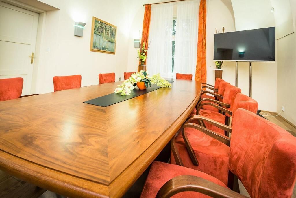 A conference room with a large wooden table, red upholstered chairs, a flat-screen TV, and a painting on the wall.