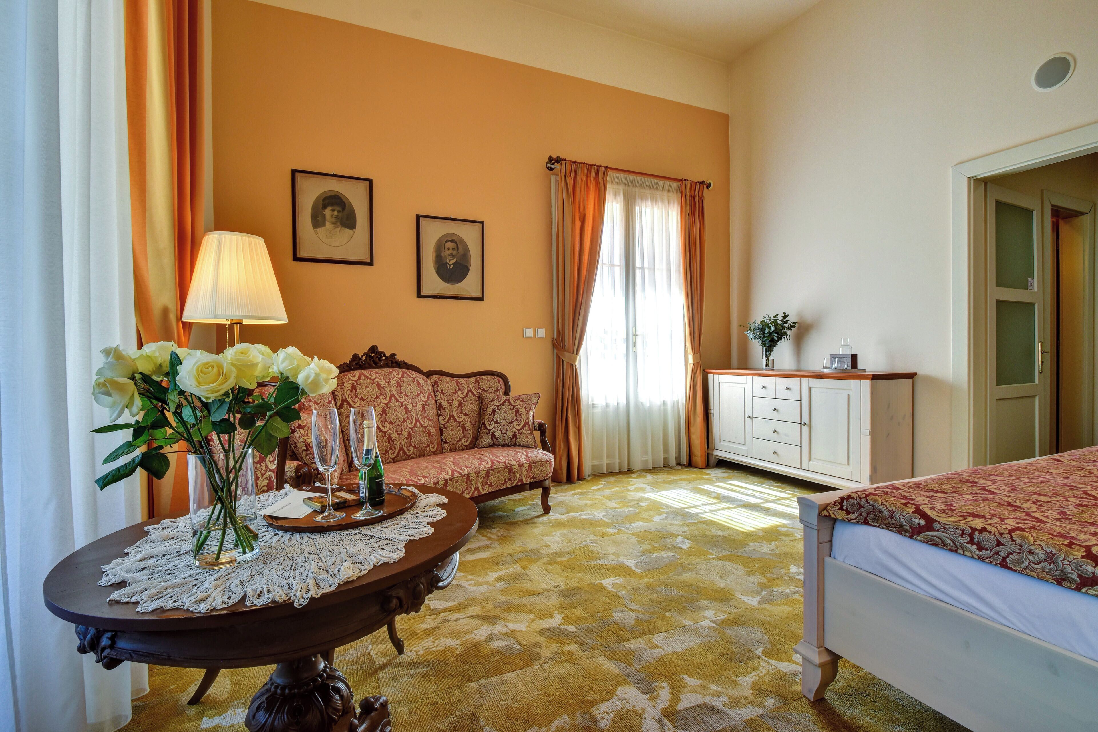 A room with a sofa, a table with a vase of flowers, a lamp, and a bed.
