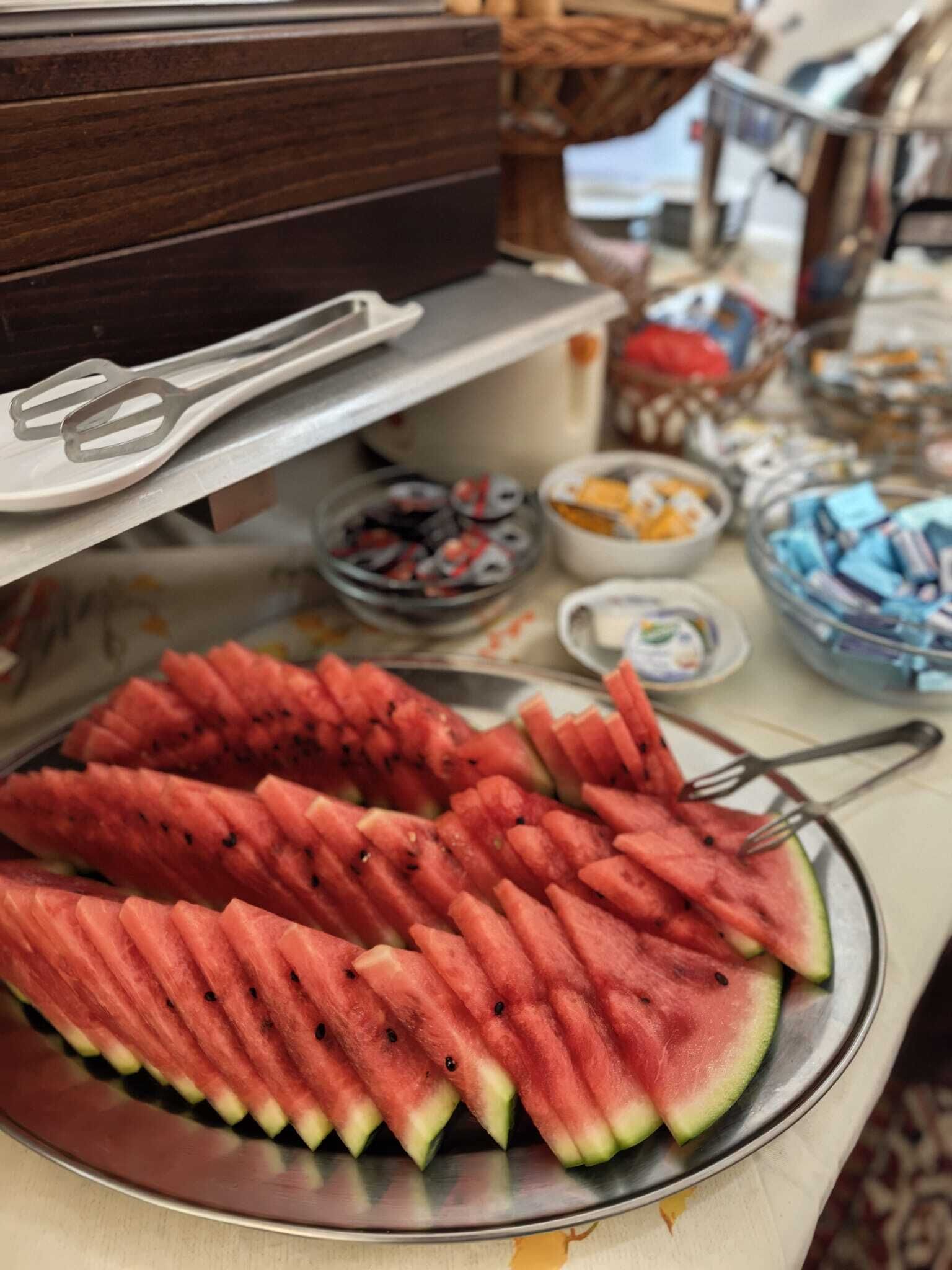Una mesa de bufé con diversas opciones de comida, incluyendo trozos de sandía, fuentes de fruta y otros bocadillos.