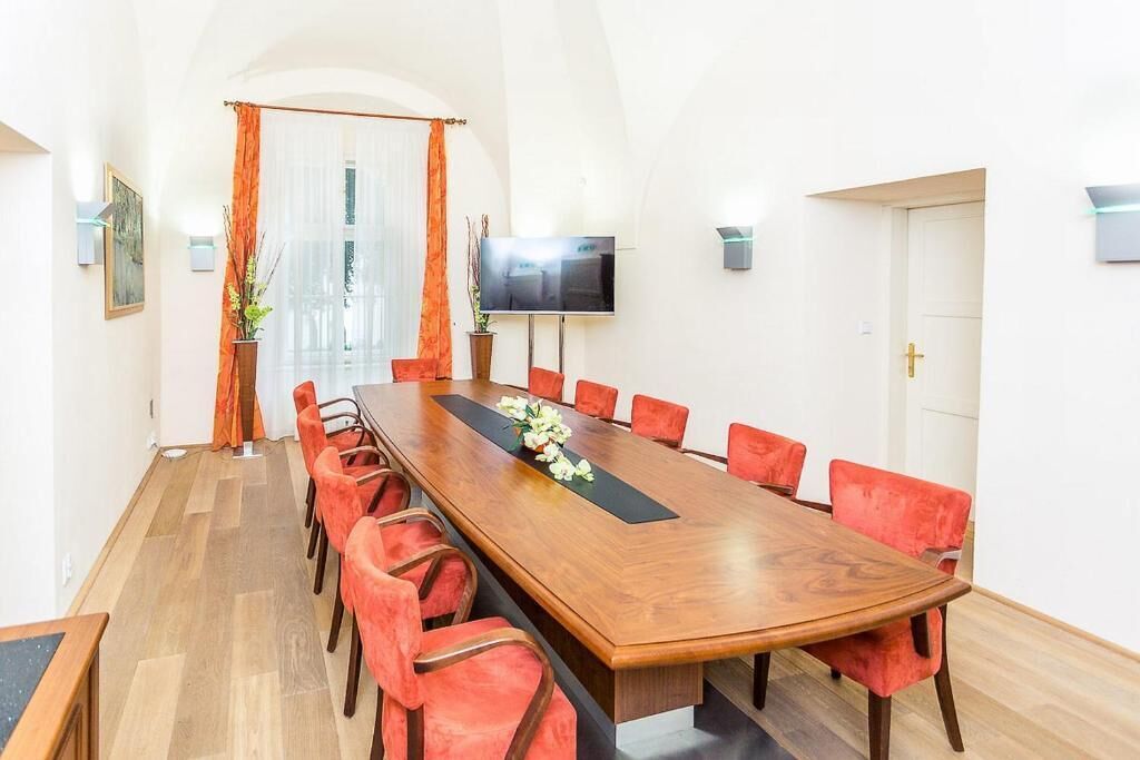 Una sala de conferències amb una taula de fusta llarga, cadires vermelles, una televisió a la paret i una porta amb una creu.
