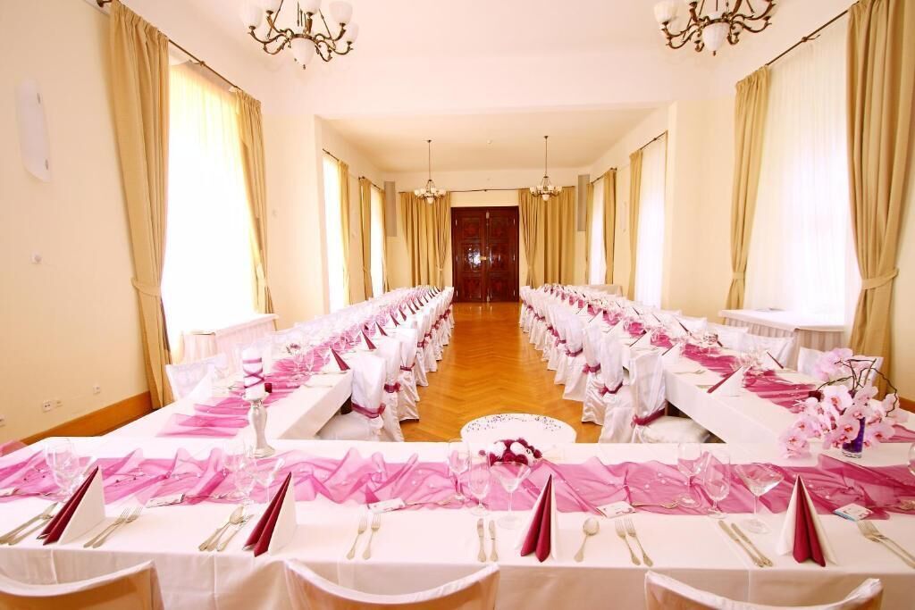 Uma sala de banquetes com longas mesas preparadas para um evento formal, com toalhas brancas e detalhes em rosa.