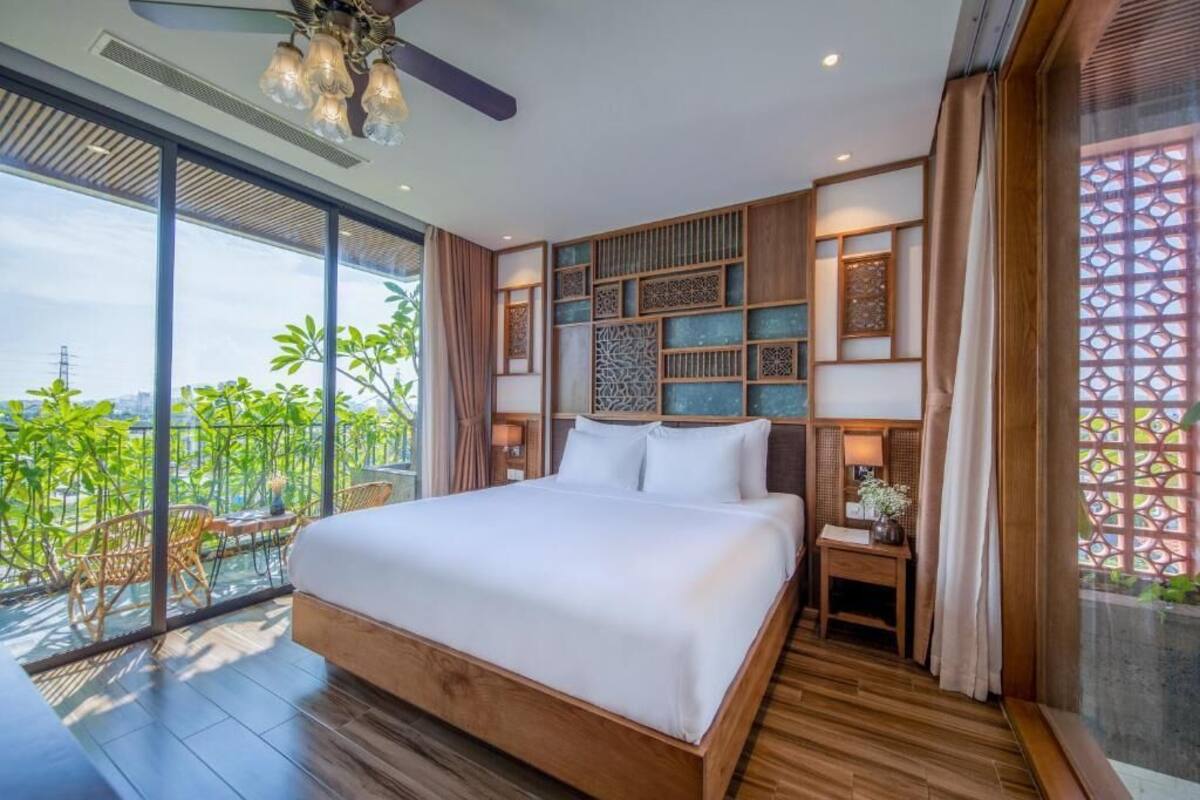 A modern bedroom with a large bed, wooden flooring, a balcony with chairs, and a view of greenery.