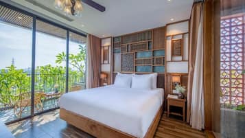 A modern bedroom with a large bed, wooden flooring, a balcony with chairs, and a view of greenery.