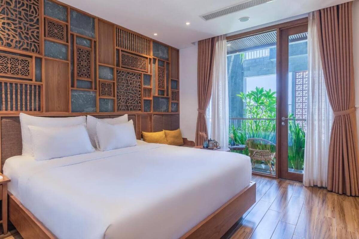 A hotel room with a large bed, wooden headboard, decorative wall panels, a balcony with curtains, and a view of greenery outside.