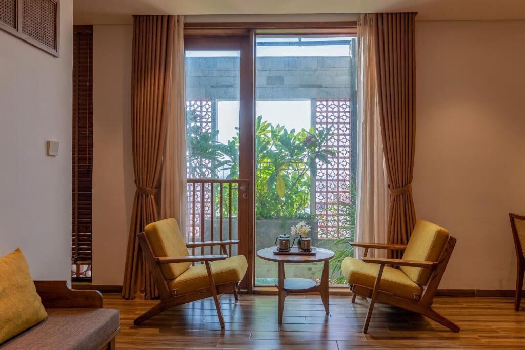 A room with two yellow armchairs, a small round table with a teapot and flowers, and a sliding glass door leading to a balcony with plants and a decorative fence.