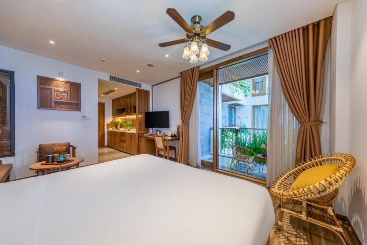 A hotel room with a large bed, a dining area, a TV, and a balcony with plants.