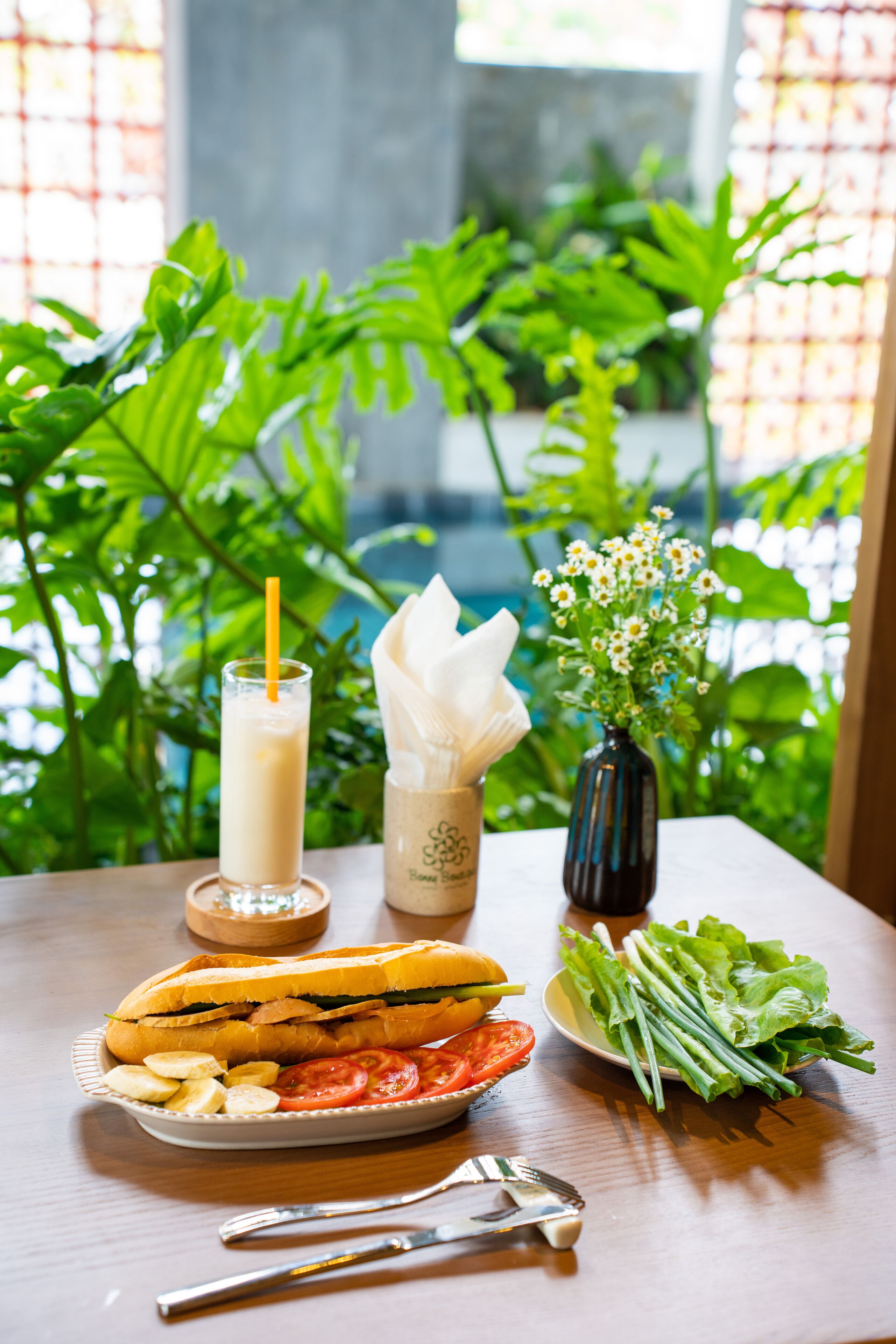 A table with a sandwich, salad, and a drink, set against a backdrop of greenery.