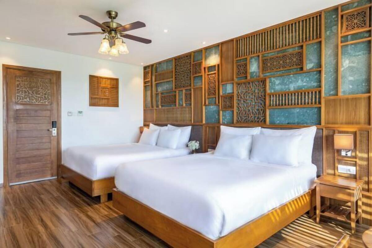 A hotel room with two beds, a ceiling fan, a decorative wall panel, and a wooden door.