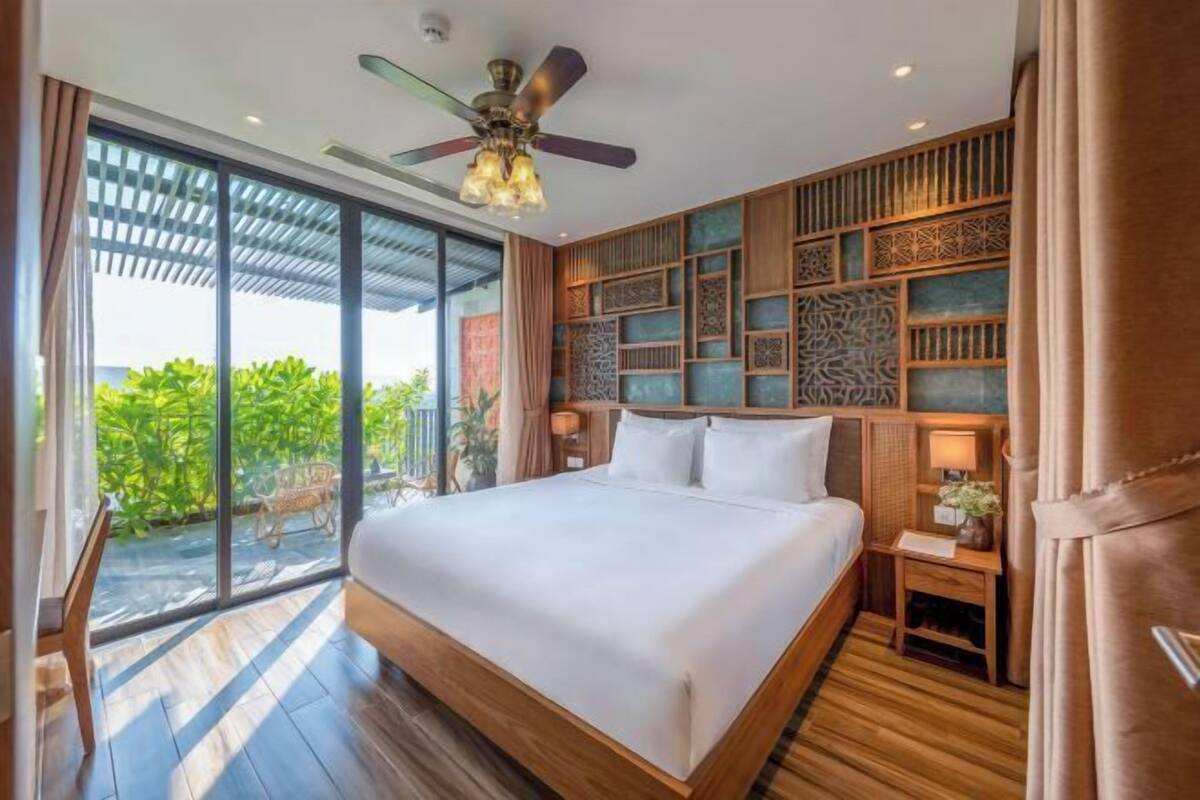 A bedroom with a large bed, a ceiling fan, a nightstand, and a view of greenery outside.