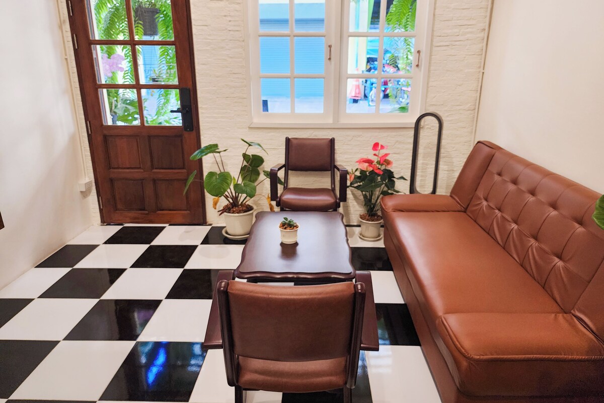 A room with a checkered floor, a brown leather sofa, two brown leather chairs, a wooden table, and potted plants.
