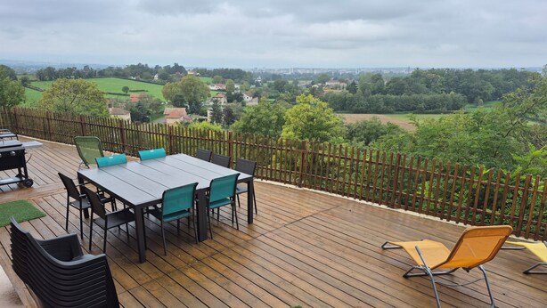 A wooden deck with a table and chairs, a grill, and a view of a green landscape. - Maison en Pierre Avec Jacuzzi été et Hiver (Perreux)
