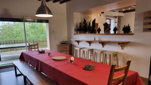 A dining area with a long table covered in a red tablecloth, wooden chairs, and a bench. There are wooden shelves above the table and a large window with a view of greenery outside. - Maison en Pierre Avec Jacuzzi été et Hiver (Perreux)