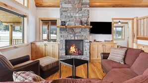A cozy living room with a stone fireplace, wooden furniture, and large windows offering a view of the outdoors.