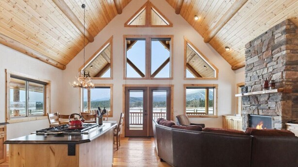 A spacious living area with a stone fireplace, wooden ceiling, and large windows offering a view of the outdoors.