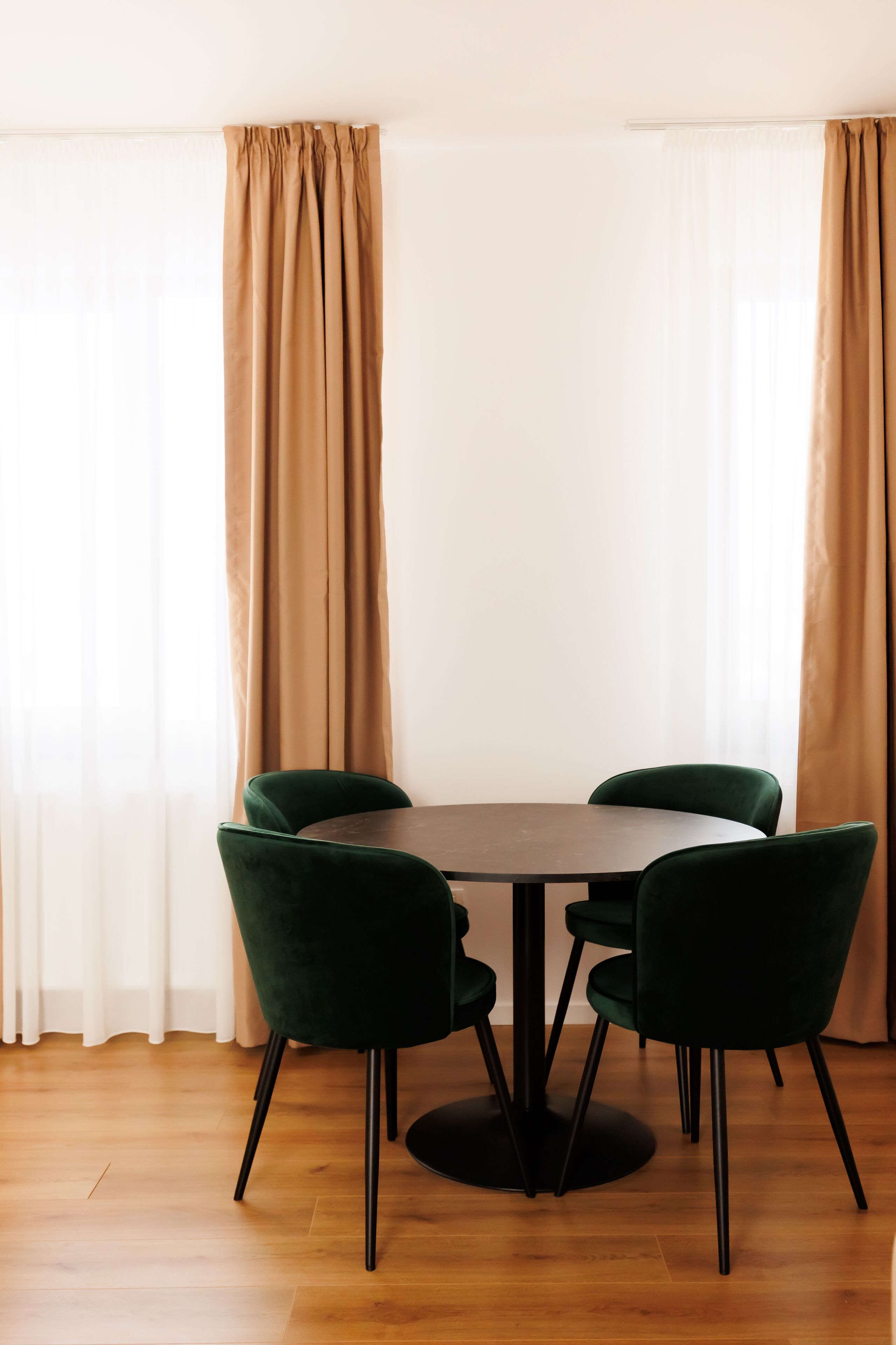 Zona pranzo con tavolo rotondo e quattro sedie verdi, circondata da tende beige e pavimento in legno.