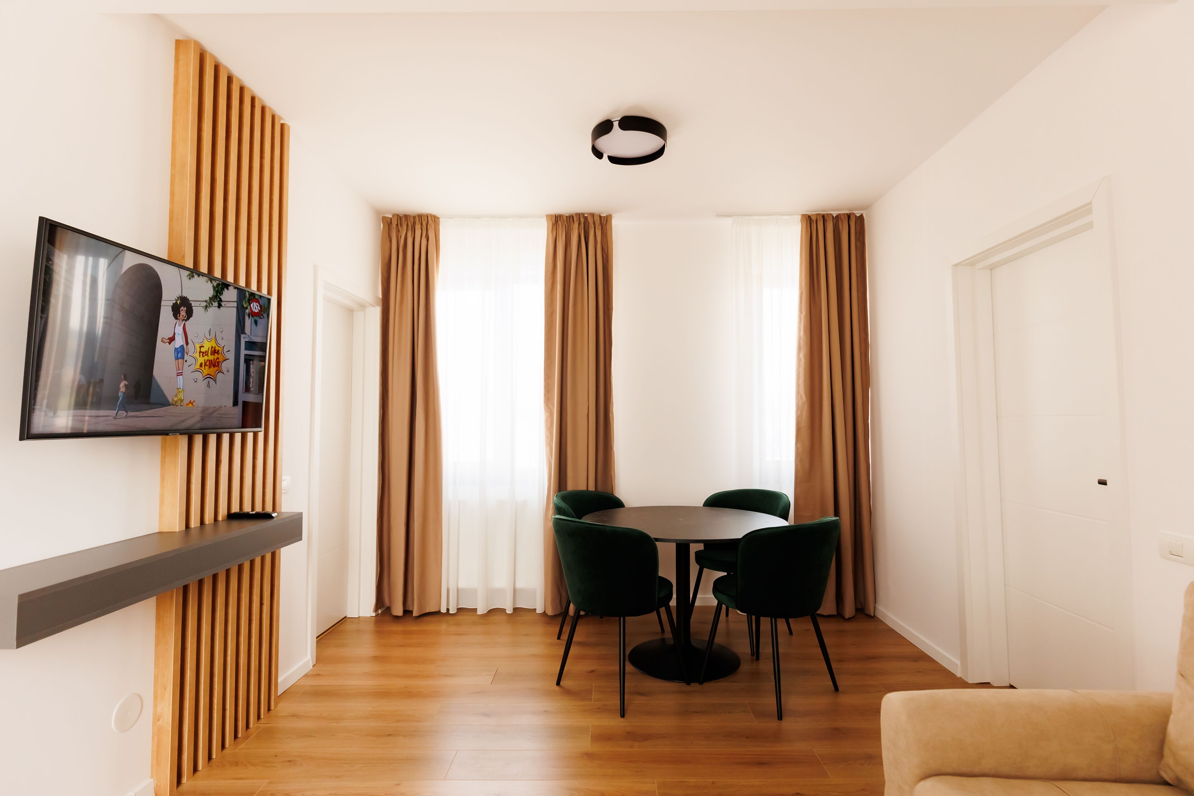 Een moderne woonkamer met een flatscreen-tv bevestigd aan een houten paneel, een ronde eettafel met vier stoelen en een beige bank.