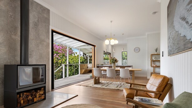 A modern living room with a fireplace, a dining area with a wooden table and chairs, and a view of the outdoors through large windows.