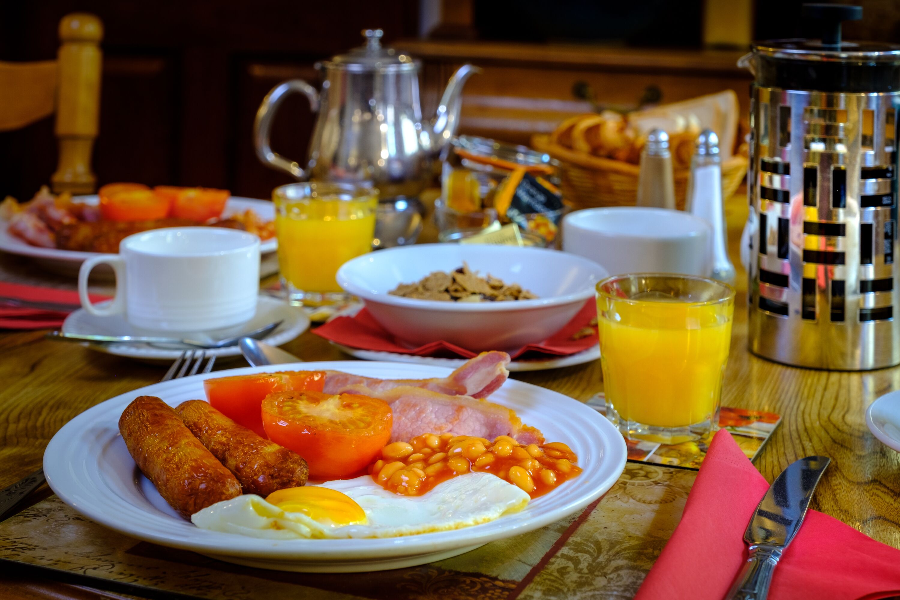 A breakfast table with plates of eggs, bacon, tomatoes, beans, and sausages, along with cups of coffee and orange juice.