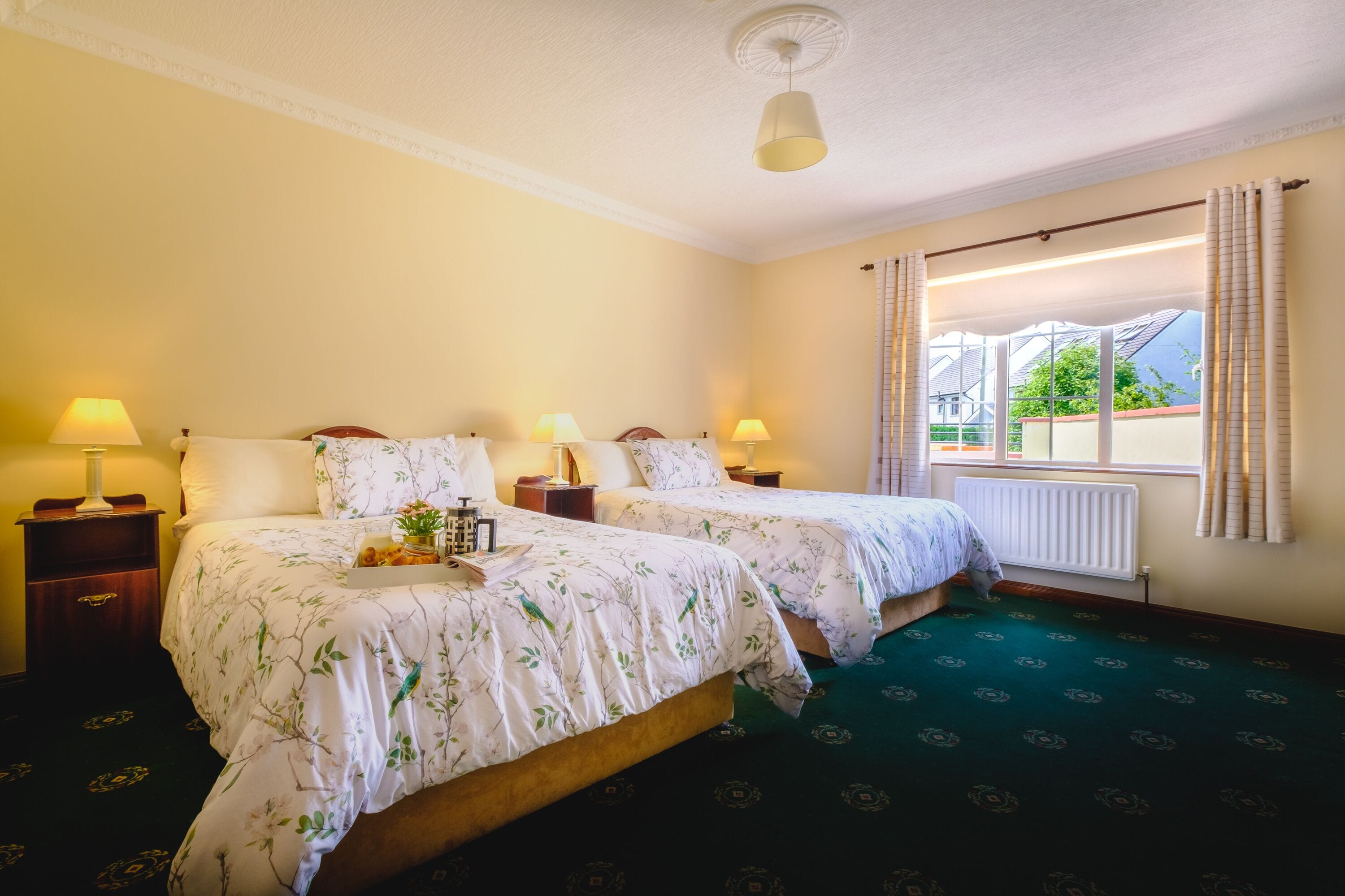 A hotel room with two beds, each with a lamp and a bedside table. There is a window with curtains and a view of the outside.