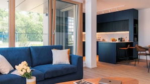 A modern living room with a blue sofa, a coffee table, and a laptop. There is a kitchen area with dark cabinetry and a bar stool. - Media Bay Apartments (Luxembourg City)
