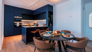 A modern kitchen with dark cabinetry, a central island, and a dining area with brown chairs and a round table. - Media Bay Apartments (Luxembourg City)