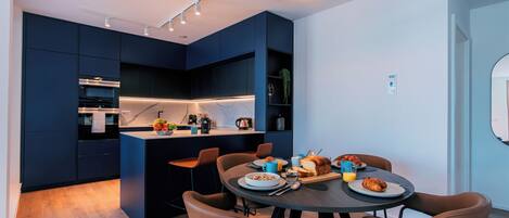 A modern kitchen with dark cabinetry, a central island, and a dining area with brown chairs and a round table.