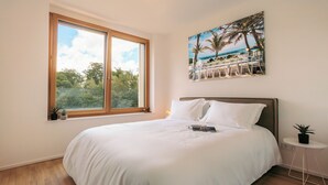 A bedroom with a large bed, a window with a view of trees and a beach, a small table with a plant, and a painting of a beach scene. - Media Bay Apartments (Luxembourg City)