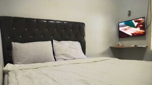 Un lit bien fait, avec une literie blanche, une tête de lit noire recouverte de tissu et un téléviseur fixé au mur, sur le côté droit.