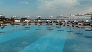 A large outdoor swimming pool with multiple lounge chairs and umbrellas, surrounded by a clear sky.