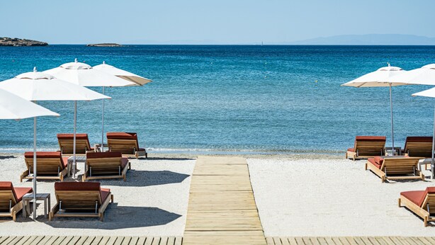A beachside view with wooden lounge chairs and white umbrellas, a wooden walkway leading to the water, and a clear blue sea. - 91 Athens Riviera, The Resort (Vari-Voula-Vouliagmeni)