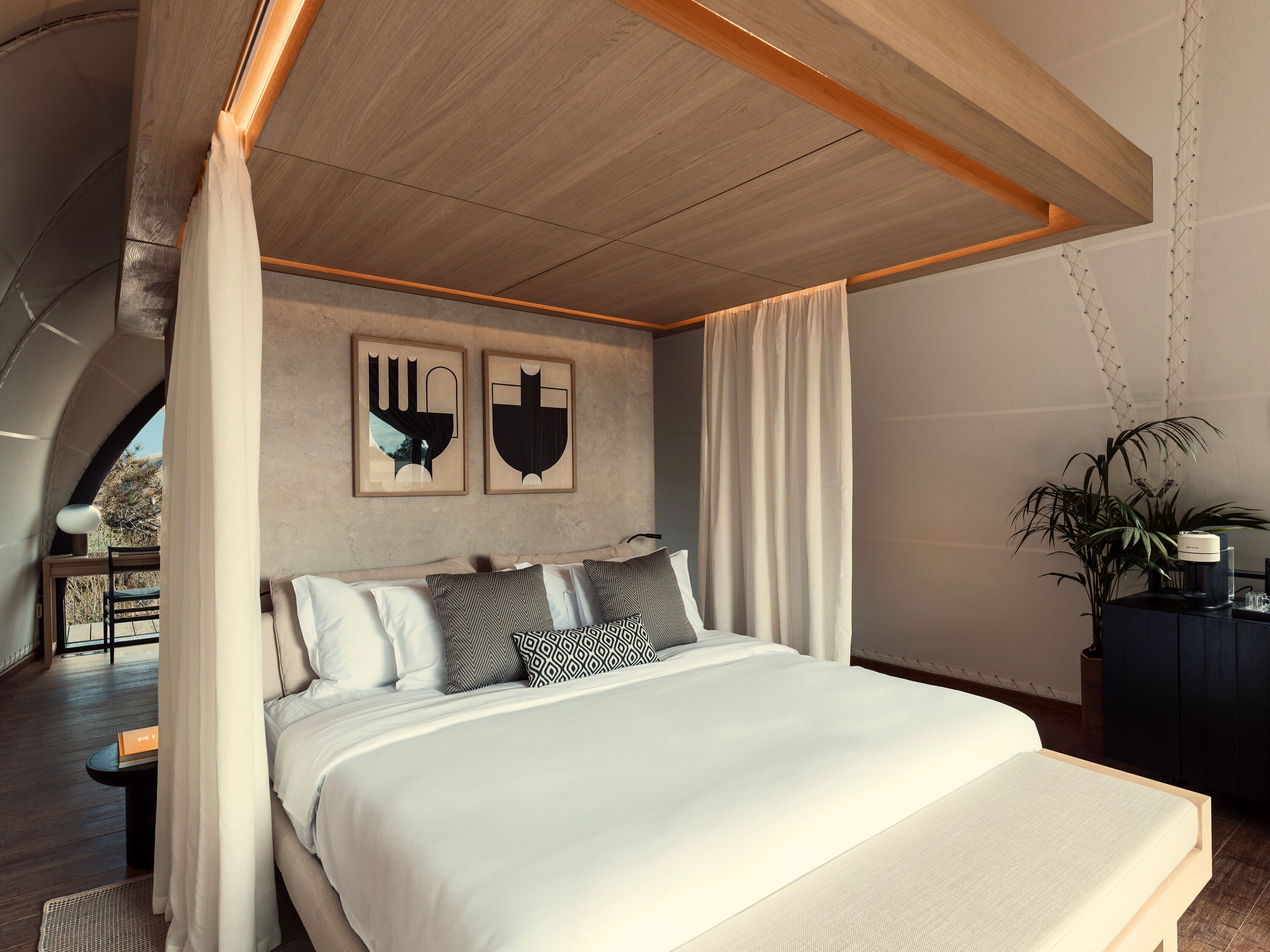 Un dormitorio moderno con una cama grande, techo con vigas de madera, dos obras de arte colgadas en la pared y una planta en maceta.