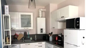 A compact kitchen with white cabinets, a washing machine, a microwave, and a hanging light fixture. - Logement 1 à 6 P- 150m Plage- Jardin et Parking (Courseulles-sur-Mer)